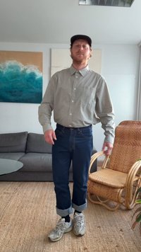 Homem com camisa de xadrez, jeans escuros arregaçados e ténis, em pé na sala de estar com uma cadeira de rattan, sofá cinza e uma pintura com tema oceânico.
