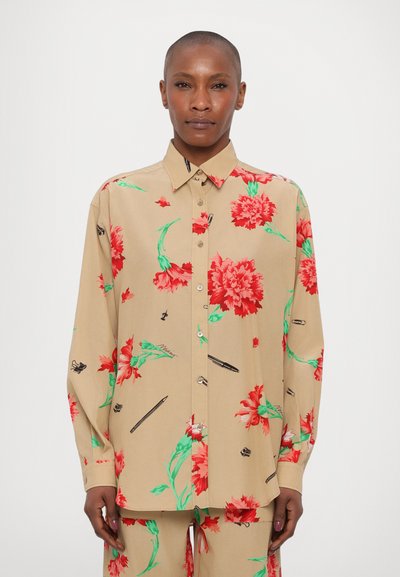 Chemise florale beige avec des motifs de fleurs rouges et vertes, fabriquée en tissu doux. Elle présente un design à boutons et des manches longues.