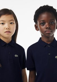 Dwoje dzieci nosi granatowe koszulki polo z teksturowanej tkaniny. Każda koszulka ma zielone logo Lacoste, a kołnierzyki są zapinane na guziki.