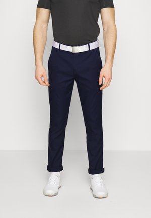 Homme portant un pantalon bleu marine, une ceinture blanche, une chemise noire et des chaussures de golf blanches, debout devant un fond uni.