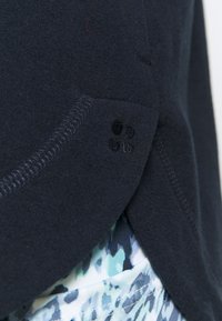 Tkanina z polaru w kolorze granatowym o fakturowanym wykończeniu, z zaokrąglonym dołem i małym czarnym logo po boku. Nałożona na wzorzystą tkaninę.