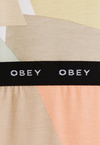 Kolorowa tkanina z wzorami, zawierająca geometryczne kształty w odcieniach beżu, zieleni i brzoskwini, z czarnym pasem, na którym znajduje się napis „OBEY” w białym kolorze.