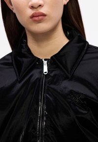 Jachetă neagră, lucioasă, cu guler, prevăzută cu închidere prin fermoar și un logo discret pe piept. Textură netedă și design ajustat.