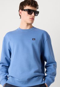 Blue cotton sweatshirt with crew neck and long sleeves. Features a small logo patch on the chest and a smooth, textured finish.