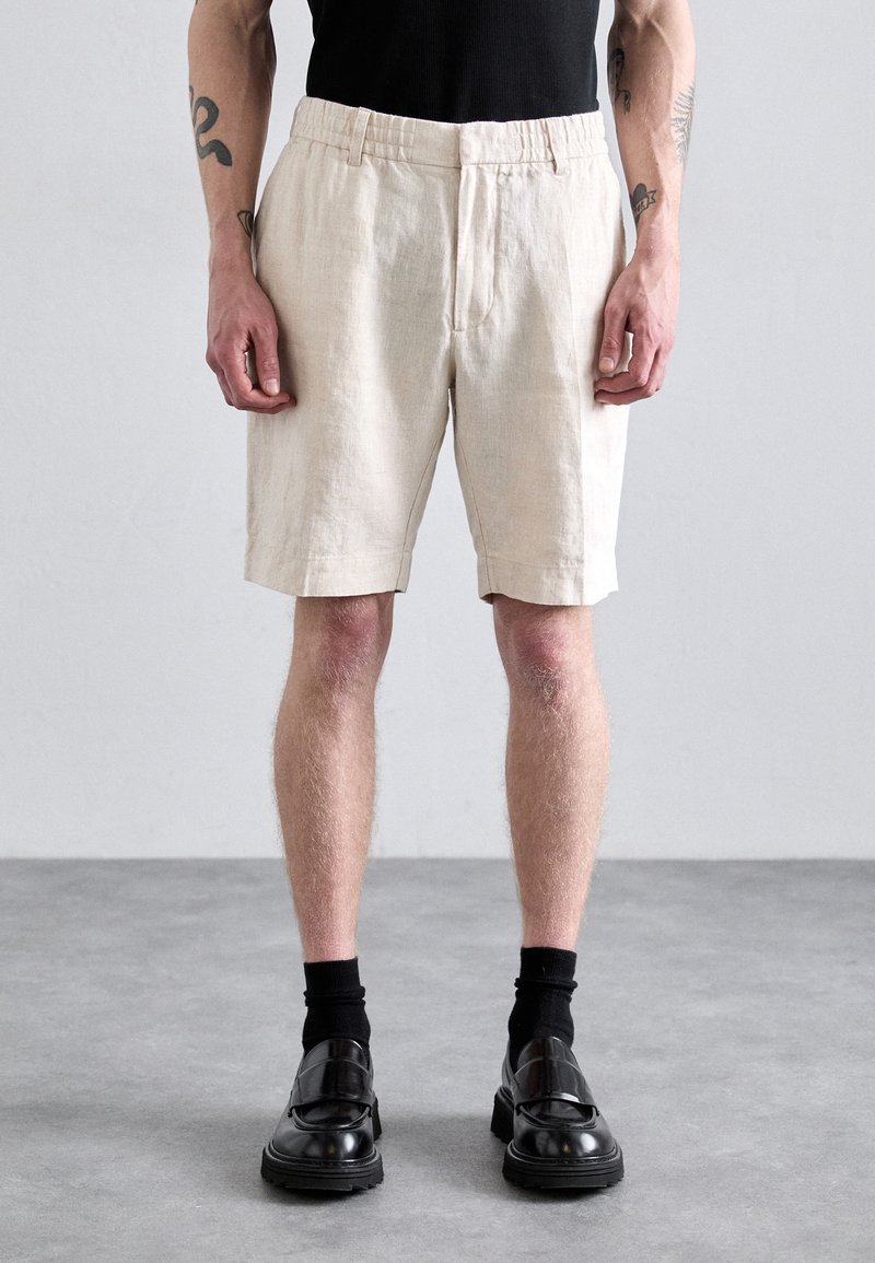 Leinen-Shorts in hellem Beige mit lässigem Schnitt, flachem Vorderteil, seitlichen Taschen und elastischem Bund, kombiniert mit schwarzen Schuhen und Socken.