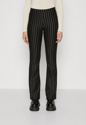 Person trägt schwarze Nadelstreifenhose, schwarze klobige Schuhe und ein langärmeliges cremefarbenes Oberteil und steht vor einer schlichten Wand.