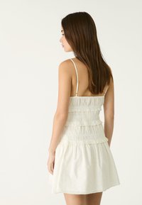 Cremfarbenes Minikleid mit verstellbaren Spaghettiträgern, gerafftem Oberteil und Rüschensäumen. Hergestellt aus leichtem Stoff mit strukturierter Oberfläche.