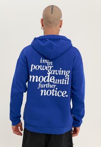 Felpa blu realizzata in morbido tessuto, con grande testo bianco sulla parte posteriore: "sono in modalità risparmio energetico fino a nuovo avviso." Cappuccio regolabile.