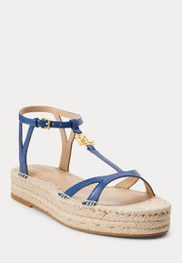 Sandale en cuir bleu avec des brides entrecroisées, un accent en logo doré et une semelle en espadrille tissée. Talon plat, design à orteils ouverts.