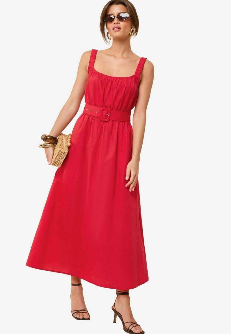 Rochie maxi roșie cu decolteu pătrat, bust plisat și curea la talie. Textură de țesătură netedă, purtată cu sandale cu bretele și accesorii.