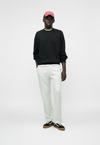 Sudadera negra, pantalones de chándal gris claro, zapatillas negras con acentos blancos y una gorra rosa. Conjunto casual con un ajuste relajado y diseño sencillo.