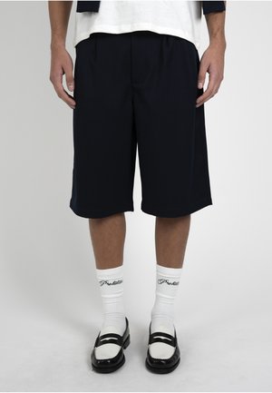 Man draagt zwarte knielange shorts, witte sokken met zwarte tekst en zwart-witte loafers en staat tegen een eenvoudige achtergrond.