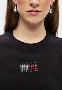 Czarna bluza z logo ozdobionym kryształami w kolorach czerwonym, białym i niebieskim; okrągły dekolt o gładkiej fakturze.