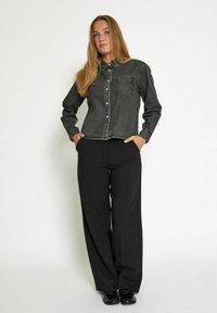 Donna con capelli biondi lunghi sta in piedi con le mani in tasca, indossando una giacca di denim nera, pantaloni larghi neri e scarpe nere, di fronte a una parete bianca.