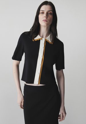 Młoda kobieta o prostych czarnych włosach, ubrana w czarną, krótkorękawową bluzkę na guziki z białym i musztardowym kołnierzykiem oraz czarną spódnicę.