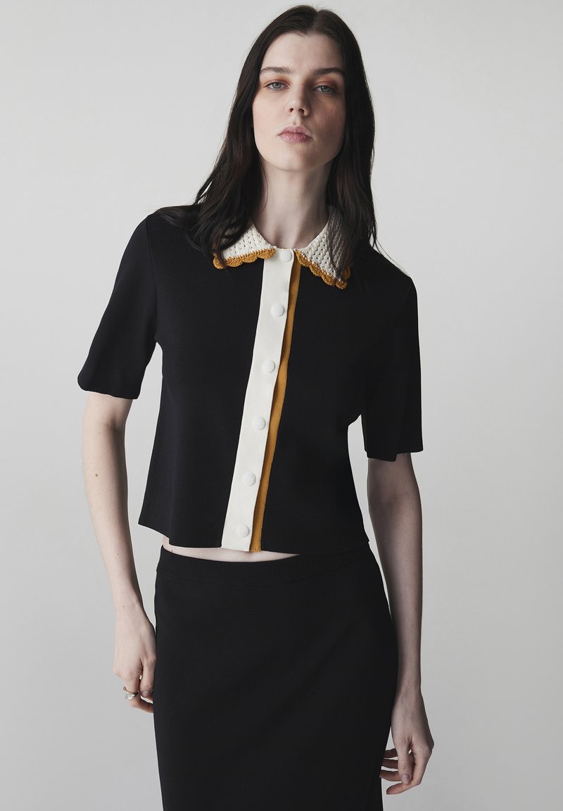 Mujer joven con cabello negro liso, que lleva una blusa negra de manga corta con botones, cuello blanco y mostaza, y falda negra.