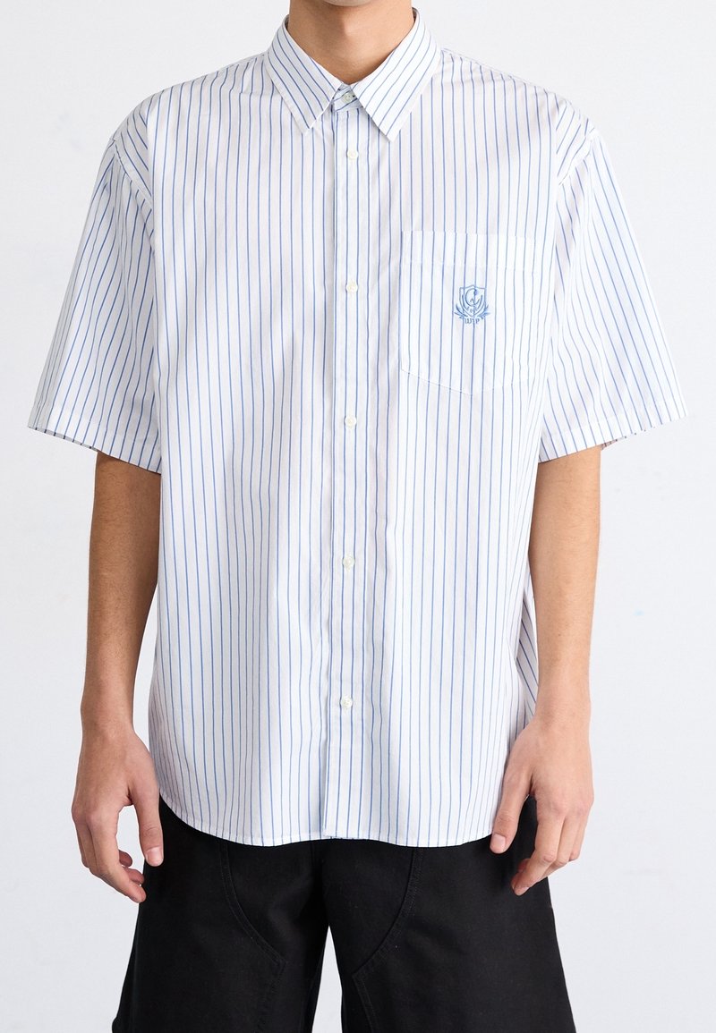 Lyhythihainen, napillinen paita valkoisesta kankaasta, jossa siniset pystysuorat raidat. Vasemmassa rinnassa tasku ja pieni kirjailtu logo.