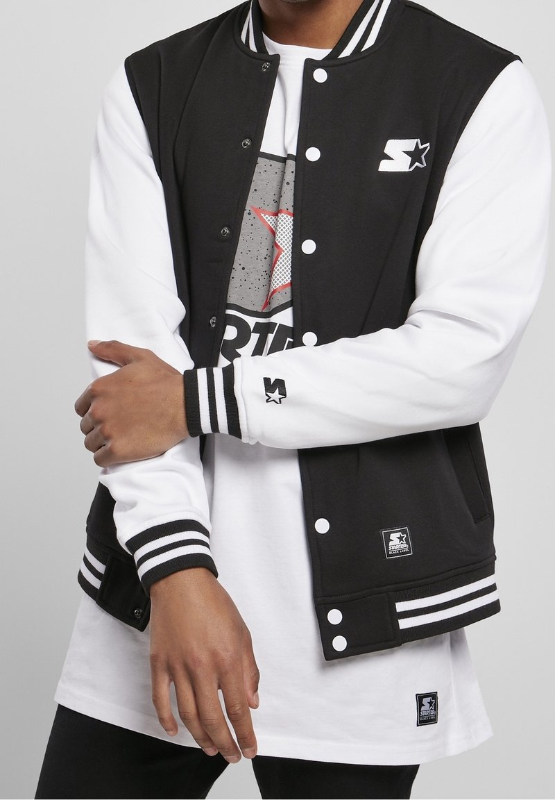Zwart-witte varsityjack met gestreepte ribgebreide manchetten, drukknoopsluiting en logo-accenten. Draagt over een grijze graphic t-shirt.