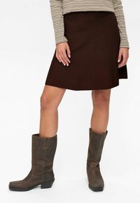 Femme portant une jupe marron jusqu'au genou, un haut à rayures à manches longues et des bottes en cuir marron foncé mi-mollet, se tenant sur un fond blanc.