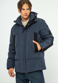 Námořnický puf z bundy s kostkovaným vzorem, kapucí, černými akcenty, předními kapsami a texturovaným povrchem. Obsahuje zip a knoflíkové zavírání.