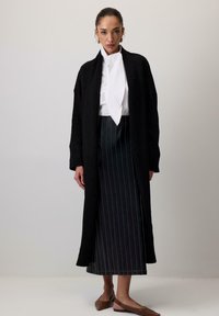 Lungo cardigan nero realizzato in tessuto testurizzato, abbinato a una blusa bianca e a una gonna a righe scure, completato da scarpe flat marroni.