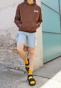 Hnědá mikina s kapucí a přední kapsou, světle modré cargo šortky, žluté ponožky a černé sandály, na pozadí texturované zdi.