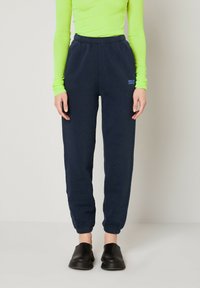 Pantalons de survêtement bleu marine avec taille élastique, poignets resserrés et détail de logo discret. Portés avec un haut à manches longues vert vif et des chaussures noires.