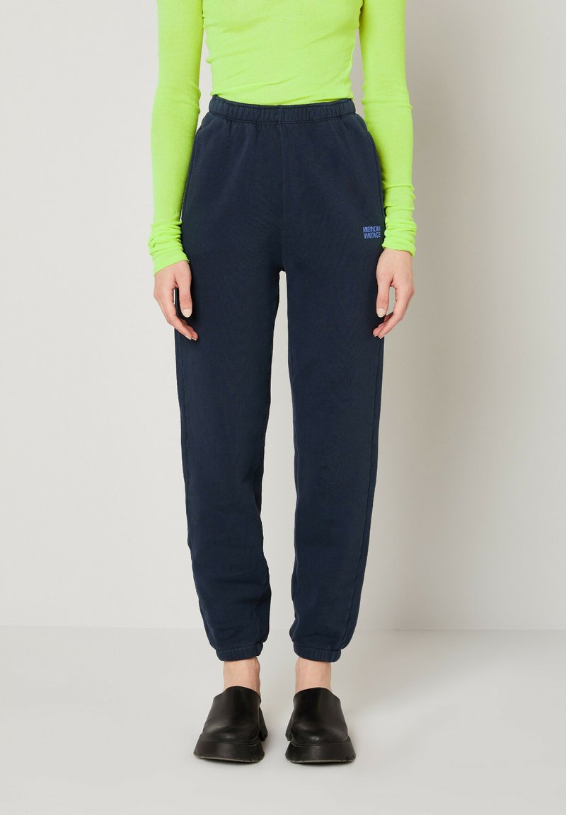 Pantalons de survêtement bleu marine avec taille élastique, poignets resserrés et détail de logo discret. Portés avec un haut à manches longues vert vif et des chaussures noires.