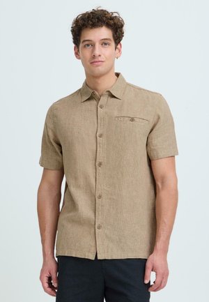 Noorus mees lokkis juustega, kandes beežikat lühikese varrukaga nööpidega särki ja tumedaid pükse, ühtlase hele taustaga.