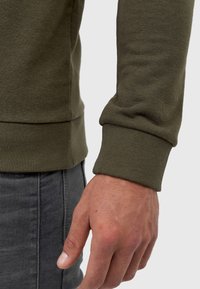 Olivengroene sweatshirtstof met geribbelde manchetten en zoom. Close-up van de pols en hand die textuur en stiksel detail tonen.