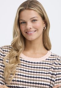 Adīta džemperis ar raksturīgu dizainu zemes toņos, tam ir rozā viļņots izgriezums un īsas piedurknes, mīksta tekstūra.