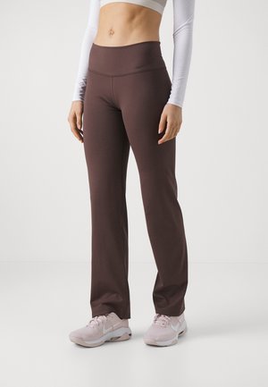 Frau trägt braune High-Waist-Yogahose, weißes langärmeliges Crop-Top und hellrosa Nike-Sneakers, steht vor einem einfarbigen Hintergrund.