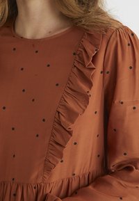 Vestido marrón con lunares negros, con un acento con volantes a lo largo del lado. Mangas largas y textura gather en la cintura.