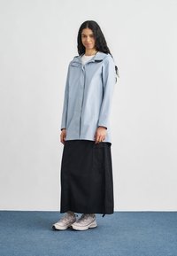 Svetlomodrá nepremokavá bunda s kapucňou, predným zipsom a bočnými vreckami. Nosená cez dlhú čiernu sukňu a svetlé tenisky.