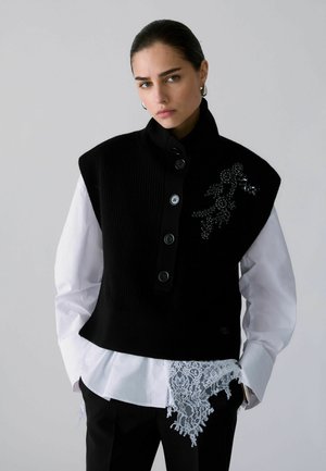Femme portant un gilet noir boutonné avec détails perlés sur une chemise blanche à bordure en dentelle, debout les mains dans les poches devant un fond gris.
