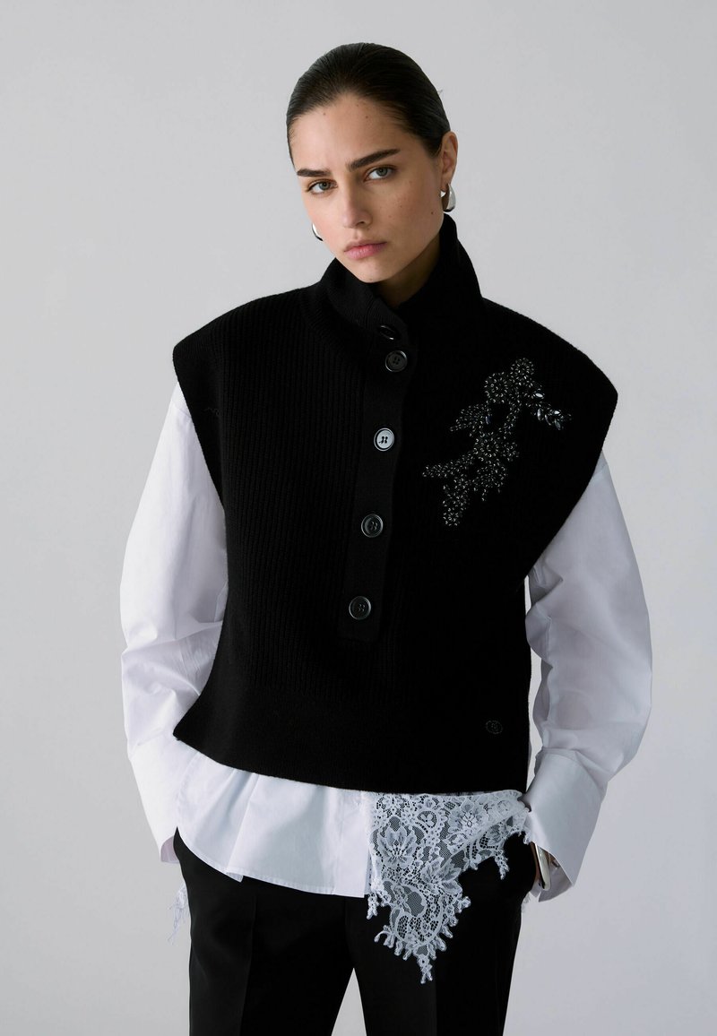 Femme portant un gilet noir boutonné avec détails perlés sur une chemise blanche à bordure en dentelle, debout les mains dans les poches devant un fond gris.