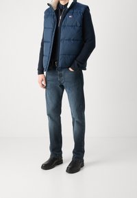 Marinblå pufferväst med krämfärgad innersida, bärs över en marinblå tröja. Matchas med blå jeans och svarta, texturerade ankelkängor.