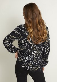 Sort bluse med et bølget hvidt og olivengrønt mønster, lange ærmer og en afslappet pasform. Har en rund kant og tekstureret stof.