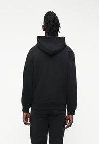 Μαύρο φούτερ με κουκούλα από ύφασμα με υφή, διαθέτει μεγάλη κουκούλα και μανσέτες με ριγέ σχέδιο. Εμφανίζεται από πίσω μαζί με σκούρο παντελόνι.