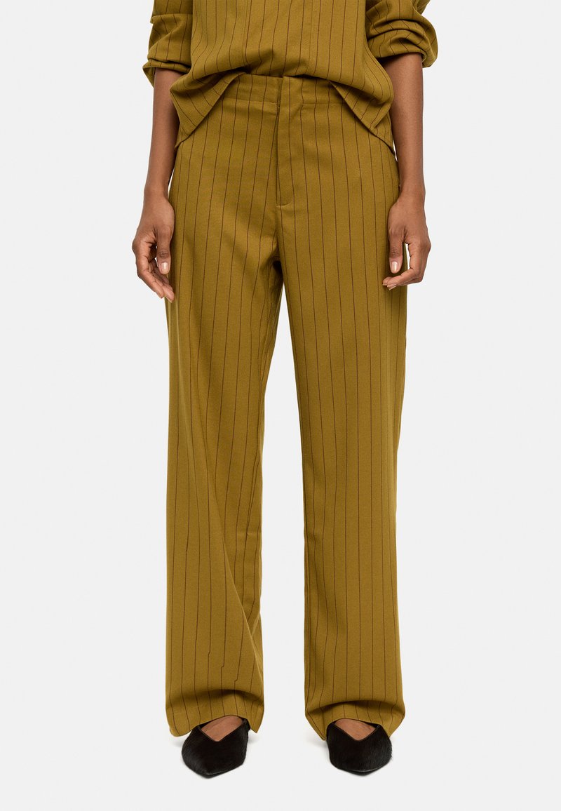 Oliwkowe spodnie z wysokim stanem w pionowe paski, o luźnym kroju i miękkiej fakturze, zestawione z czarnymi szpilkami.