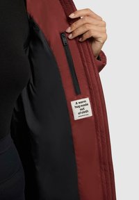 Bordeaux geïsoleerde jas met een zwarte voering, voorzien van een zijzak met rits en een stoffen label met de tekst: "Een warme knuffel gemaakt van stof."