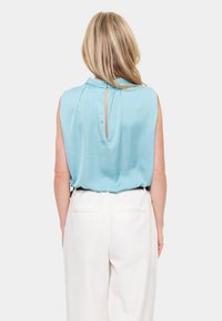 Blusa senza maniche di un azzurro chiaro con dettaglio a chiave sul retro e scollatura plissettata, abbinata a pantaloni bianchi a gamba larga. Tessuto dalla texture liscia.