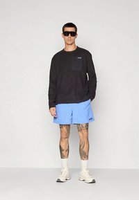 Moški, oblečen v črno majico z dolgimi rokavi, svetlo modre kratke hlače, bele nogavice, bele superge, temna sončna očala, stoji z eno roko v žepu na enobarvnem ozadju.