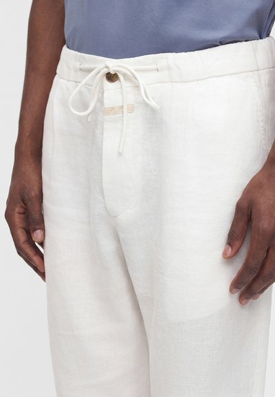 Gros plan d'une personne portant un pantalon en lin blanc avec une taille à cordon et un bouton, et une chemise bleue rentrée.