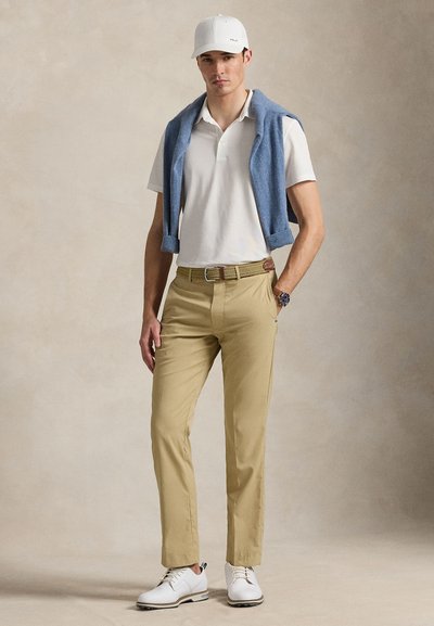 Πόλο μπλουζάκι σε λευκό, χακί παντελόνι, ανοιχτό μπλε πουλόβερ περασμένο στους ώμους, λευκά παπούτσια και λευκό καπέλο με μινιμαλιστικό σχέδιο.