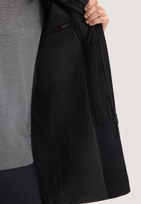 Giubbotto nero con interno imbottito e esterno liscio, dotato di una tasca con cerniera e contrasto di texture del tessuto. Maglione grigio sotto.