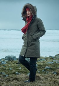 Oliven grøn parka med pelsforet hætte, rød tørklæde og strikket hue; sorte jeans og støvler; står på en klippefyldt kystlinje mod en grå himmel.