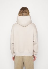 Personne aux longs cheveux portant un sweat à capuche beige clair et un pantalon couleur sable, debout dos à la caméra.
