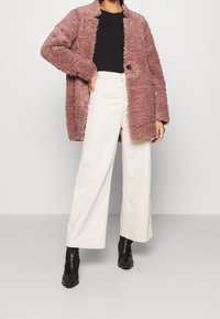 Løs, mauvefarvet yderbeklædning i blødt materiale med en enkelt knaplukning. Kombineret med hvide, højtaljede bukser med vide ben og sorte ankelstøvler.