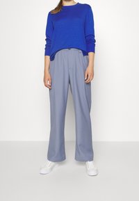 Blå tröja, lätt tyg, avslappnad passform. Ljust grå byxor, vida ben, elastisk midja. Vita sneakers fulländar outfiten.